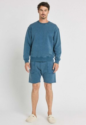 Homme debout portant un sweat-shirt bleu à manches longues, un short bleu assorti avec des poches à l'avant et des chaussures beige slip-on sur un fond blanc.