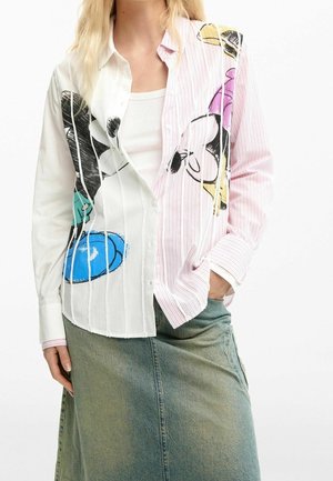 Femme portant une chemise boutonnée bicolore avec des imprimés de dessins animés sur un débardeur blanc, associée à une jupe en jean délavée.