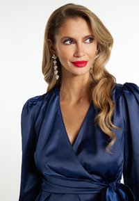Abito a portafoglio in satin blu navy con maniche a palloncino; presenta un profondo scollo a V e un dettaglio in vita annodato. Il modello indossa orecchini a goccia floreali e rossetto rosso.