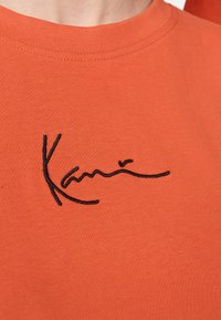 Orange bomulds t-shirt med rund halsudskæring, der har et sort broderet signaturlogo på brystområdet. Glat tekstur, afslappet design.