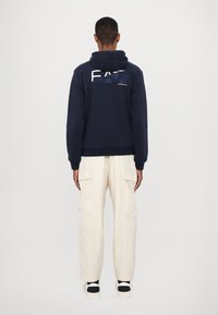 Jūrų korpuso džemperis su priekine grafika, derinamas su šviesiais bežiniais cargo ilgais šortais su šoninėmis kišenėmis. Aprangą papildo juodi ir balti sportbačiai.