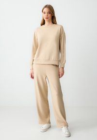 Felpa beige e pantaloni a gamba larga in morbido tessuto, con scollatura rotonda e polsini a coste. L'outfit è completato da sneaker bianche.