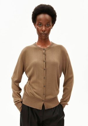 Schwarze Frau mit kurzen lockigen Haaren, trägt einen braunen, geknöpften Cardigan und schwarze Hosen, steht mit den Händen in den Taschen.
