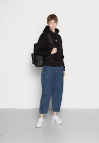 Felpa nera con cappuccio, jeans blu a vita alta con cerniere laterali, sneakers bianche e uno zaino nero. Design semplice, vestibilità casual.