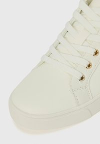 White leather sneaker with a smooth finish, round toe, and textured panels. Features white laces and gold-tone eyelets.