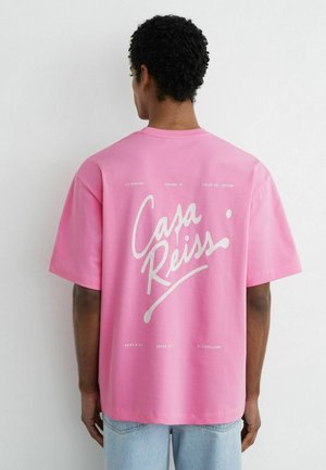 Homme portant un t-shirt rose ample avec le texte blanc « Casa Reiss » et un jean bleu clair décontracté, debout contre un mur blanc uni.