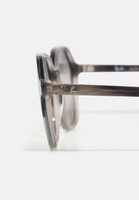 Óculos de sol com tonalidade cinza e armação transparente, apresentando um formato retangular e um logo metálico da Ray-Ban na haste.