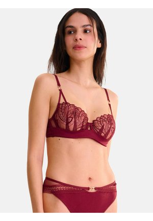 Femme portant un ensemble de lingerie en dentelle bordeaux avec des bretelles réglables et un détail décoratif doré sur la culotte, se tenant devant un fond uni.