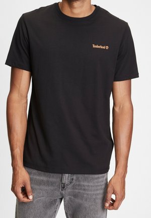 Mężczyzna noszący prostą czarną koszulkę Timberland z krótkim rękawem i szare dżinsy, stojący na białym tle.