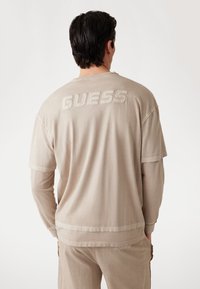 Béžové oversized tričko vyrobené z mäkkého materiálu, s embosovaným nápisom "GUESS" na zadnej strane. Má voľný strih a krátke rukávy.