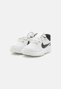 Baskets blanches avec texture côtelée, accent Swoosh noir de Nike, sangle ajustable et semelle rembourrée conçue pour le confort.