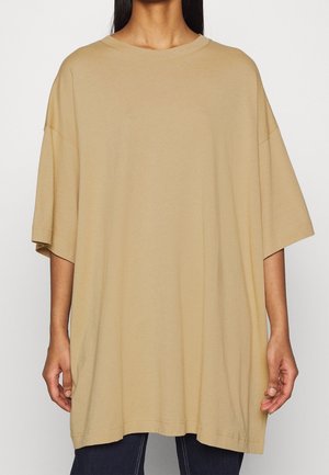 Osoba ubrana w oversize'owy beżowy T-shirt z krótkim rękawem i okrągłym dekoltem, w połączeniu z ciemnymi spodniami, na jednolitym jasnym tle.