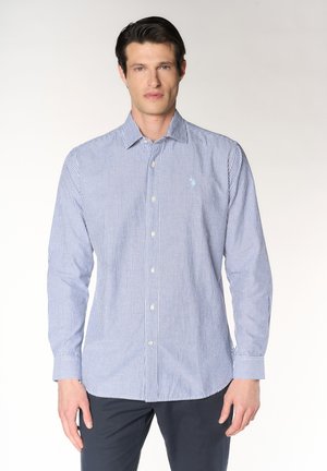 Uomo con indosso una camicia a maniche lunghe a righe verticali blu e bianche abbottonata e pantaloni scuri su uno sfondo semplice.