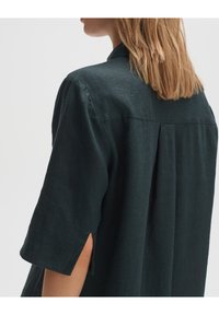 Donkergroene linnen blouse met kraag, korte mouwen en zijslitsen. De achterkant heeft een geplooid ontwerp voor extra textuur.