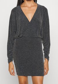 Svart klänning med djup V-ringning, långa ärmar och figursydd siluett. Tyget har en strukturerad glittrig finish med metalliska detaljer.