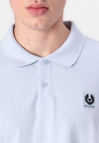Gaiši zils polo krekls ar teksturētu audumu, dubulto pogu slēgšanu un melnu logo uzlīmi kreisajā krūtī. Apkakle ir plakana.