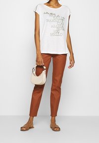 White T-shirt with multicolored text, cap sleeves. Brown fitted pants, nude sandals, and a small beige handbag with a curved handle.