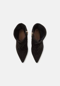 Botas de tobillo de ante negro con puntas afiladas, decoradas con tachuelas plateadas y un detalle de cremallera lateral en tono dorado. Parte superior texturizada y forro suave.