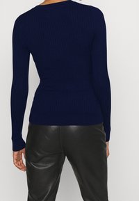 Maglione blu a coste con maniche lunghe, design aderente e scollatura rotonda. Indossato con pantaloni in pelle neri, che evidenziano la texture del capo.
