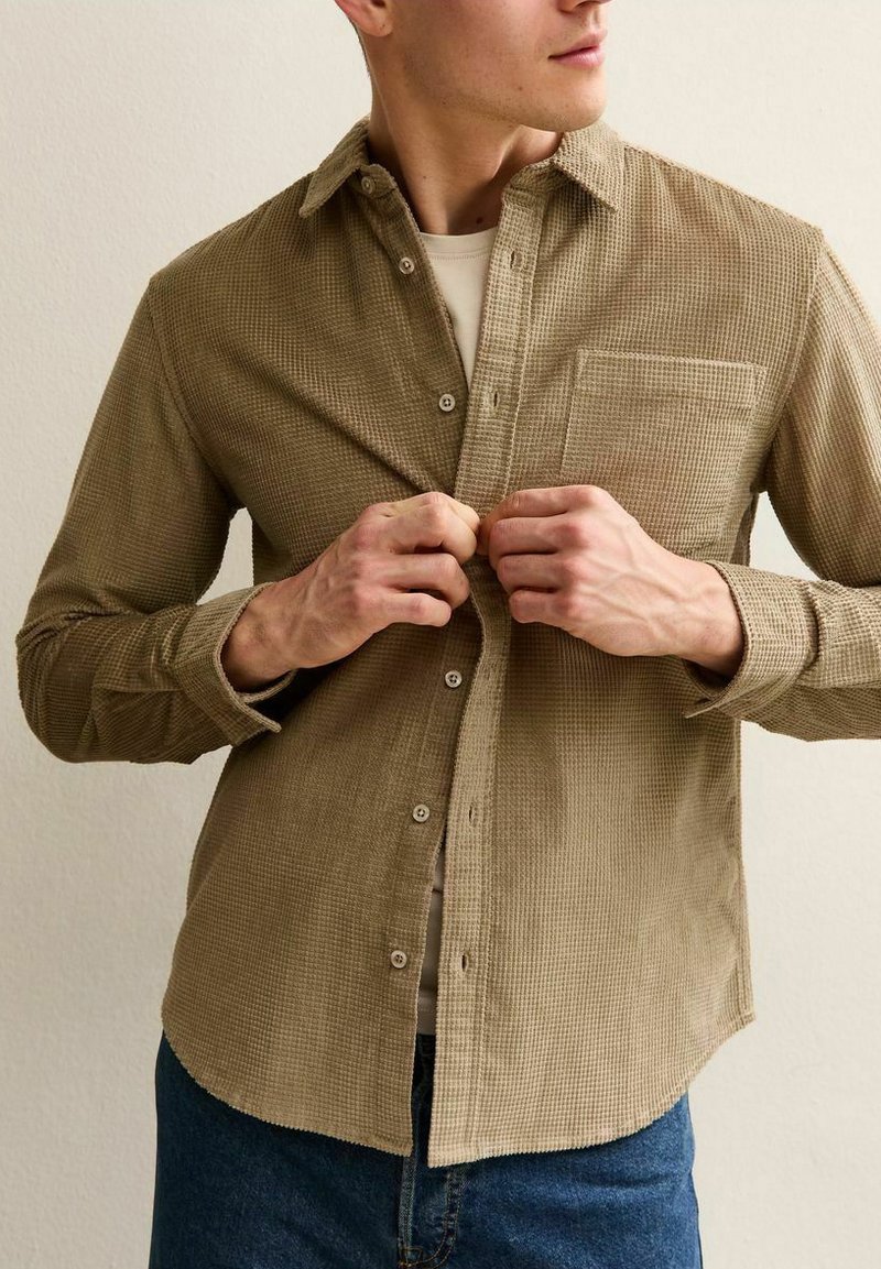 Homme portant une chemise beige texturée à boutons par-dessus un haut blanc, en train de boutonner, avec un jean bleu, devant un mur uni.