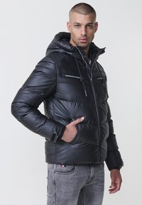 Sort pufferjakke med hætte, der har lynlåslommer, en quiltet design og en glat tekstur. Båret over grå jeans.