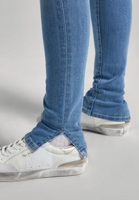 Vaqueros ajustados de color azul claro con detalle de dobladillo deshilachado, combinados con zapatillas blancas que presentan una parte superior texturizada y sutiles acentos dorados.