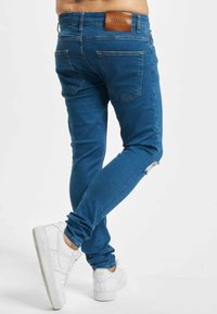 Férfi slim-fit kék farmert visel, amelyen kis szakadás van a jobb térdnél, és fehér Nike Air sportcipőt hord, háttérben fehér fal.