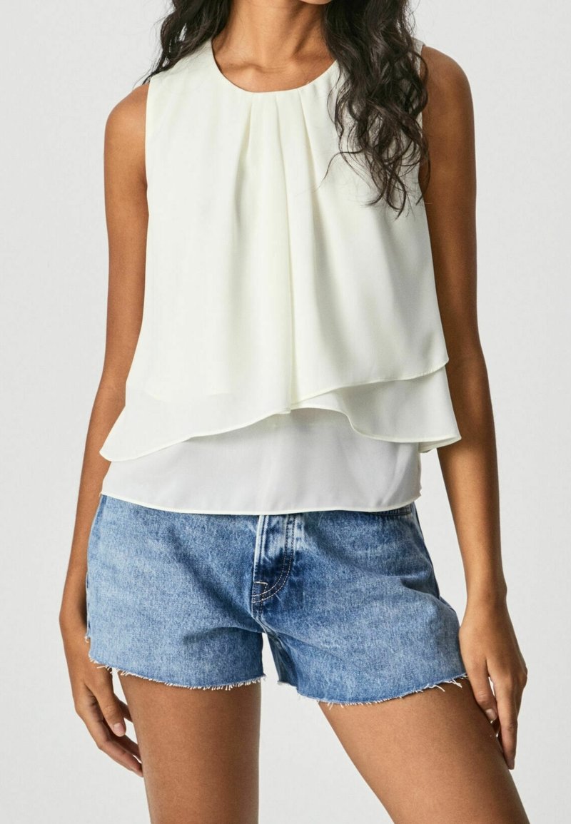 Femme portant un chemisier blanc sans manches à volants et plis, associé à un short en denim bleu clair aux bords effilochés.
