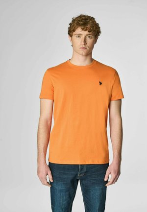 T-shirt arancione a maniche corte in cotone, con colletto girocollo e un piccolo logo nero sul lato sinistro del petto. Indossato con jeans blu.