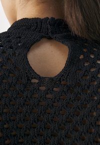 Svart strikket topp med åpen ryggdesign og sirkulært utskjæring. Det teksturerte mønsteret er intrikat og viser det vevde materialet.