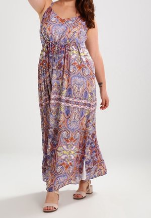 Vrouw draagt een mouwloze maxi-jurk met een multikleuren paisley patroon, een zijsplit en beige sandalen met blokhak.