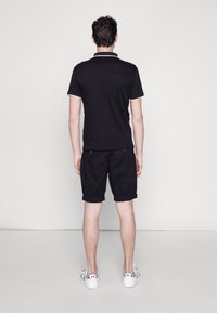 Svart pikétröja med vita krage detaljer, kombinerad med svarta shorts. Vita sneakers med rutiga detaljer. Bakifrån mot en neutral bakgrund.