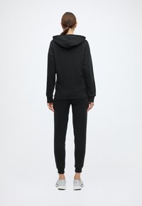 Czarna bluza z kapturem i dopasowane spodnie dresowe. Materiał jest gładki, z prążkowanymi mankietami. Noszone z jasno szarymi butami sportowymi.