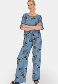 Niebieski komplet bluzki i spodni w wzory z czarnymi ptakami i liśćmi, o luźnym kroju i miękkiej tkaninie. Całość dopełniają sandały.