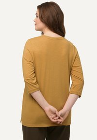 Camisa mostaza de manga tres cuartos con un cuello redondo y un corte holgado, con un dobladillo que es ligeramente más largo en la parte posterior.