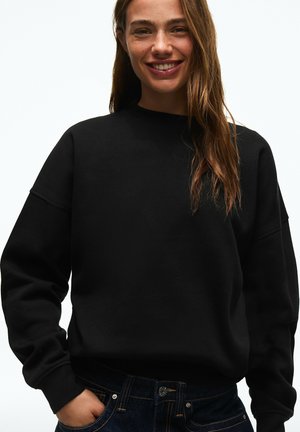 Ung kvinde med langt brunt hår smilende, iført en sort sweatshirt og mørke jeans, stående med den ene hånd i lommen.