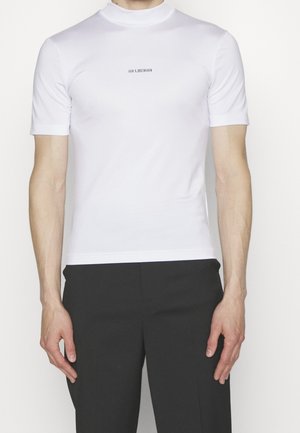 Mann trägt ein eng anliegendes weißes Kurzarmhemd mit dem Schriftzug "HAN KJØBENHAVN" auf der Brust und schwarze Hosen vor neutralem Hintergrund.