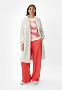 Witte textuur jas over een rood gestreepte trui, gecombineerd met feloranje wijde broek en lichte schoenen, alles tegen een neutrale achtergrond.
