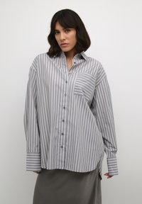 Chemise à manches longues rayée grise et blanche avec fermeture à boutons et poche poitrine. Présente des rayures contrastées aux poignets et un ourlet courbé.