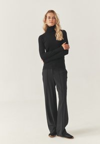 Schwarzer Rollkragenpullover mit langen Ärmeln, kombiniert mit hoch taillierten schwarzen Hosen. Glatte Stoffqualität, oben eng anliegend, unten ausgestellt.