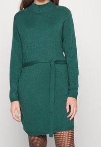 Abito in maglia a maniche lunghe di colore verde scuro con collo alto e cintura annodata in vita. Tessuto texturizzato, polsini e orlo a coste.