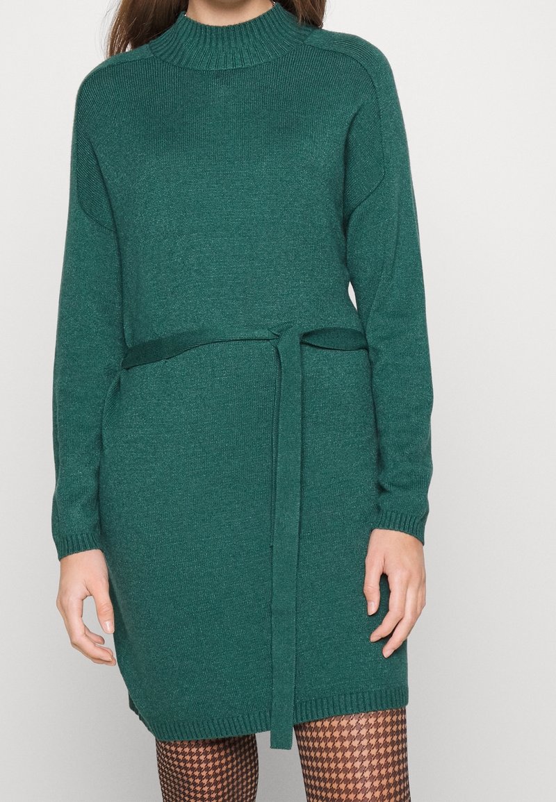 Abito in maglia a maniche lunghe di colore verde scuro con collo alto e cintura annodata in vita. Tessuto texturizzato, polsini e orlo a coste.