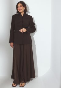 Hnedý strihaný blejzer s vysokým golierom a dlhými rukávmi, skombinovaný s prúžkovanou, plisovanou hnedou sukňou. Hladká látka s jemným leskom.