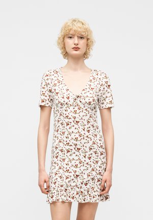 Mladi oseba s kratkimi kodrastimi blond lasmi, oblečena v belo cvetlično kratko obleko z nabranim robom, na nevtralni podlagi.