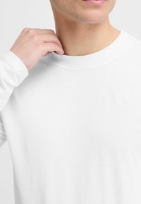 Weißes Langarmshirt mit rundem Halsausschnitt, ausgestattet mit einem dezenten, geprägten Logo auf der linken Brust und einer glatten Baumwolltextur.