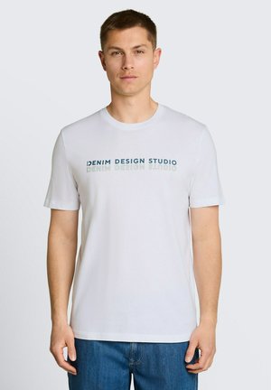 Mężczyzna w białej koszulce z napisem „Denim Design Studio” i niebieskich dżinsach, stojący na jednolitym jasnoszarym tle.