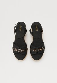 Schwarze Denim-Sandalen mit einem geknoteten Design, verstellbarem Knöchelriemen und bronzefarbenen Schnallenakzenten. Texturiertes Fußbett und juteähnliche Sohle.