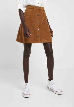Personne portant une jupe en velours côtelé marron boutonnée devant, des baskets blanches montantes et une chemise blanche aux manches retroussées, debout sur un fond uni.