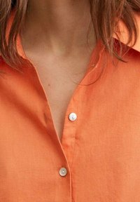 Personne portant une chemise orange à boutons blancs, col ouvert, cheveux bruns mi-longs visibles.