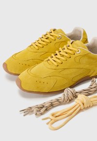 Geltoni suede sportbačiai su perforuotais detalėmis, apvaliais pirštais ir plokščiomis raištelėmis. Komplekte yra dvi raištelių poros: vieni geltoni, kiti su raštu.